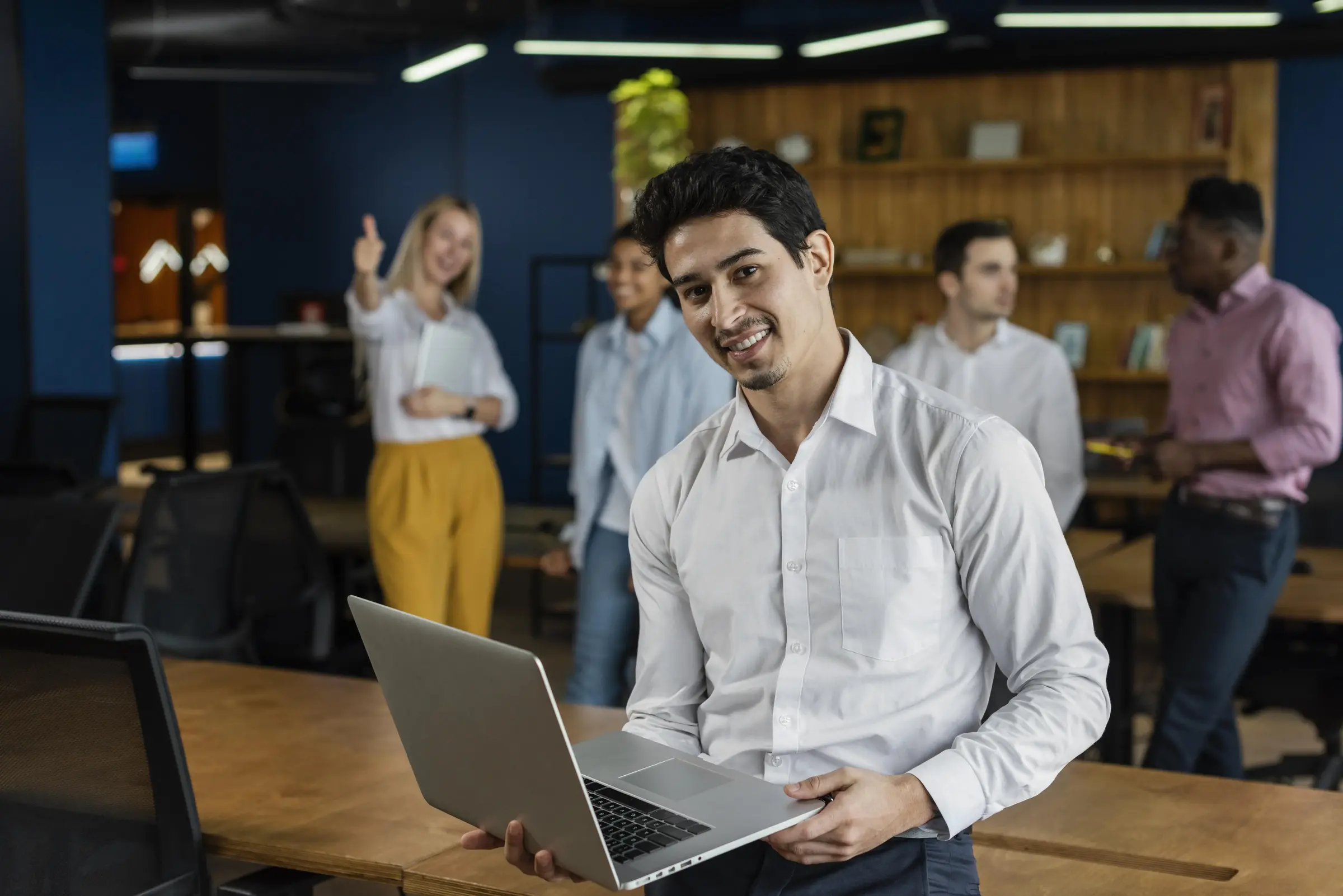 A smiling man in a white shirt holds a laptop in a modern office. Behind him, four colleagues are interacting, creating a positive, collaborative atmosphere.