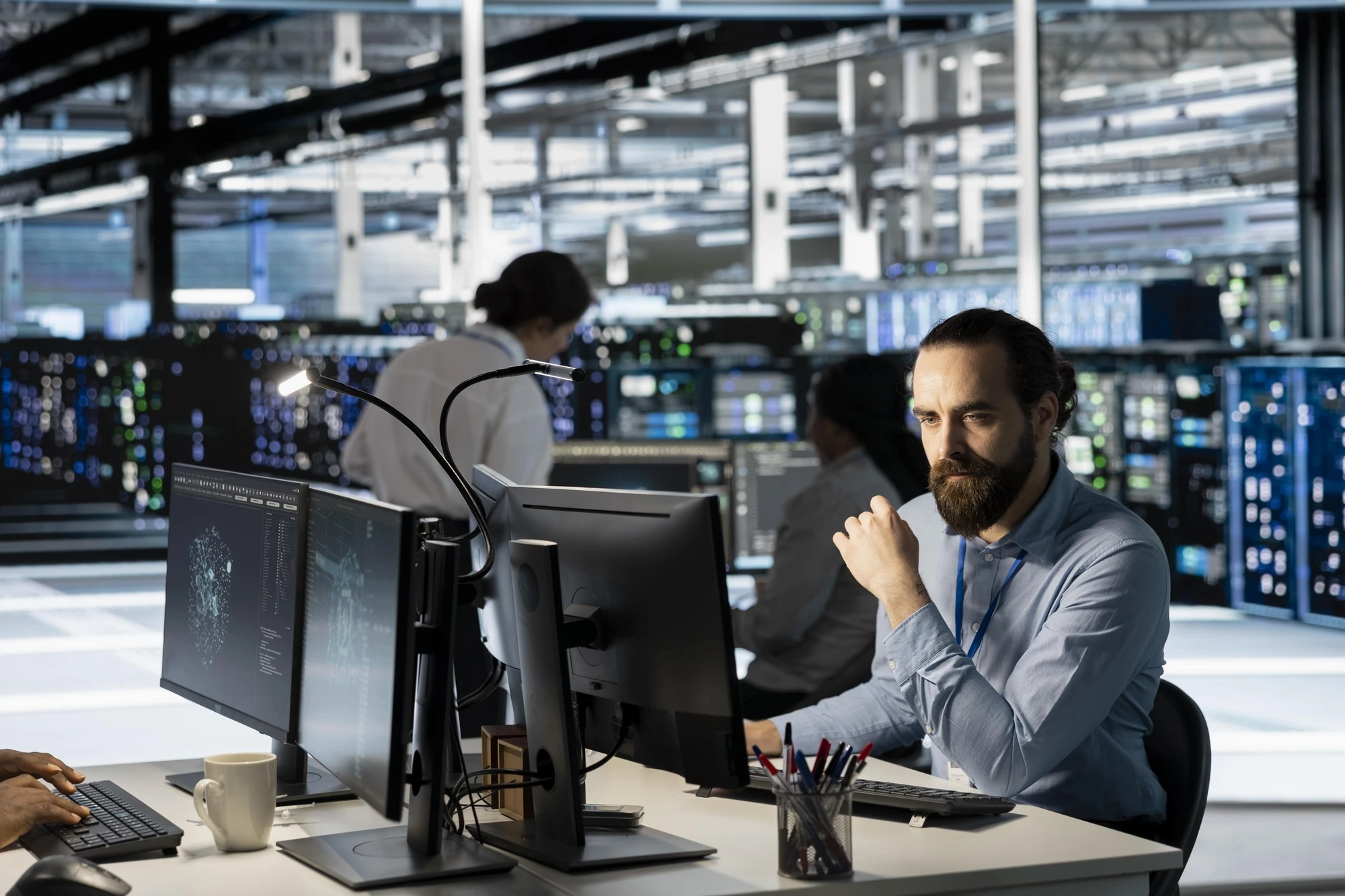 A focused man in a blue shirt works at a computer desk in a modern server room. Multiple monitors display data. The mood is focused and professional