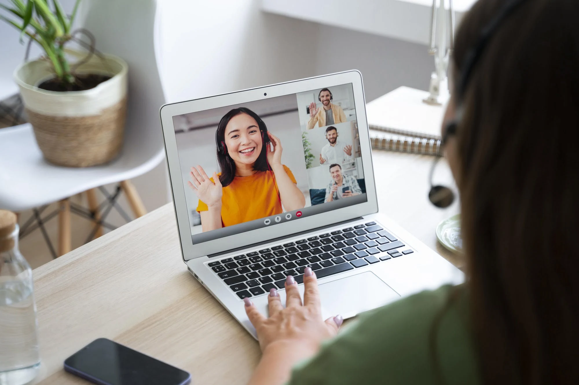 A person in a headset uses a laptop for a video call. Four cheerful people appear on screen, waving. A plant and a notebook are on the desk