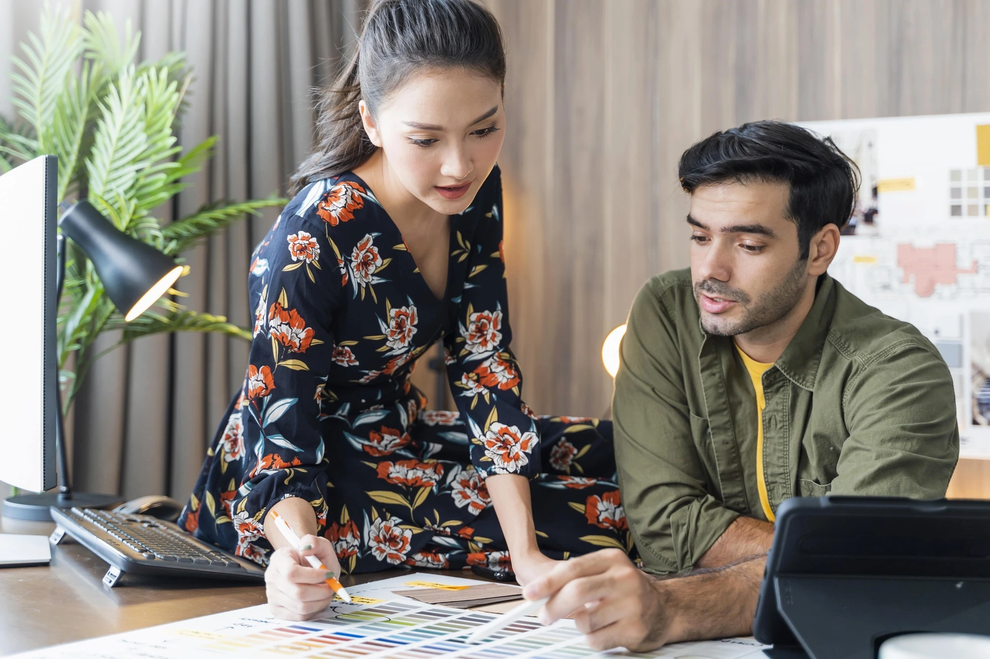A woman in a floral dress and a man in a green shirt discuss color swatches at a desk, surrounded by a computer, lamp, and plants, conveying creativity