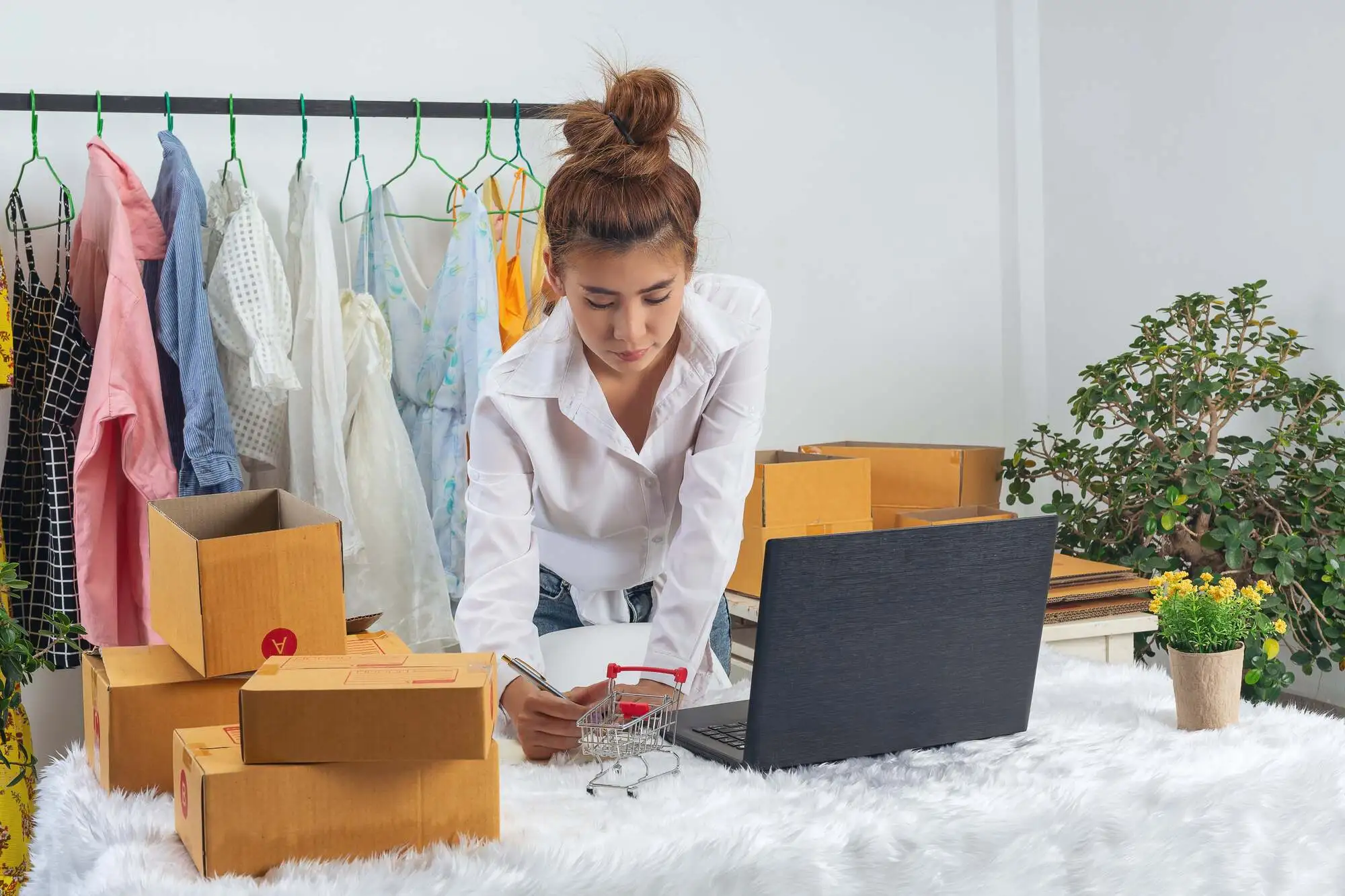 A woman in a white shirt leans over a table with boxes, writing beside a laptop. Behind her are colorful clothes on hangers. The scene conveys a focused and busy workspace