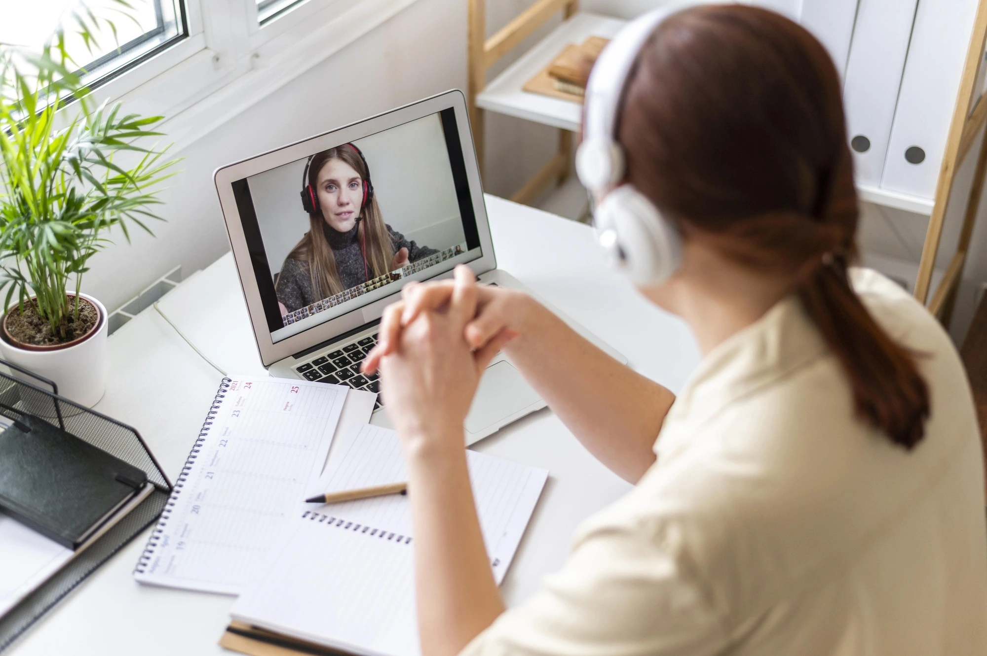 A woman wearing headphones engages in a video call on a laptop, with a notepad and pen on the desk. A plant and bright window create a calm workspace
