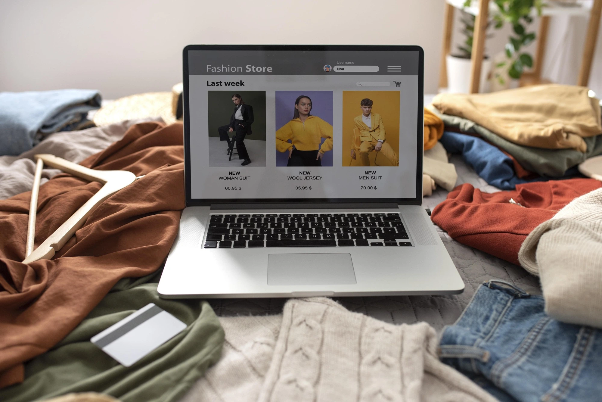 Laptop displaying an online fashion store is surrounded by casual clothes and a credit card. The atmosphere suggests online shopping at home