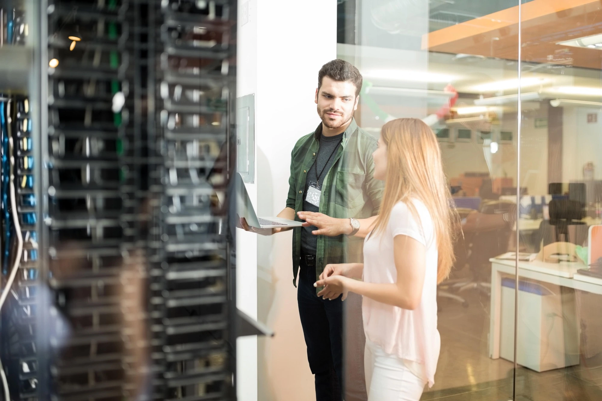 Two individuals are discussing in a server room. One holds a laptop while pointing at a server rack. The setting appears professional and focused