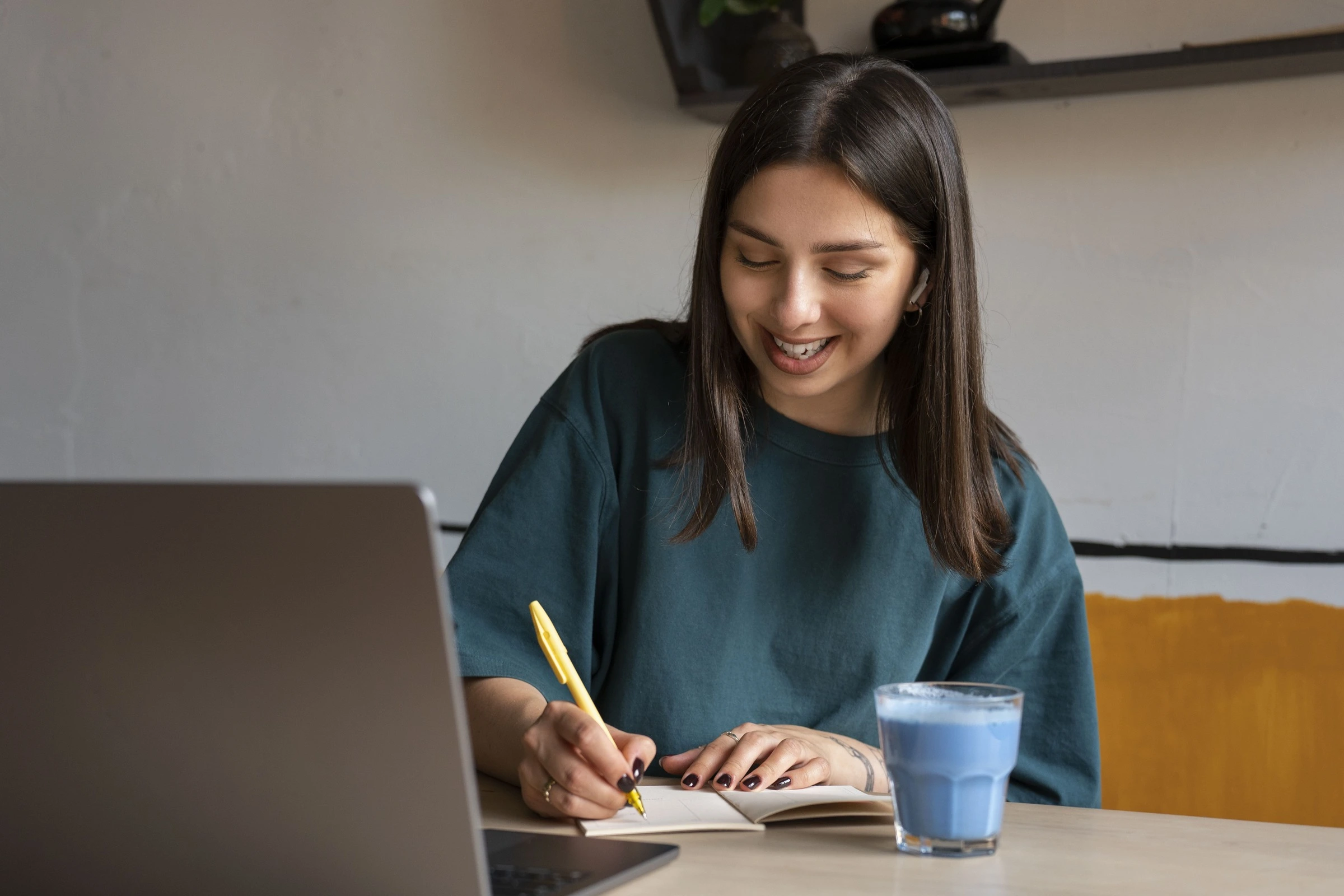 Young woman smiling while writing in a notebook at a desk with a laptop and a glass of blue milk