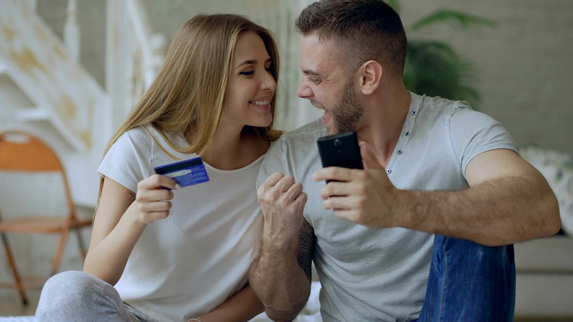 A happy couple sits together on a couch, holding a credit card and a smartphone. They gaze at each other, smiling, conveying a joyful and relaxed mood